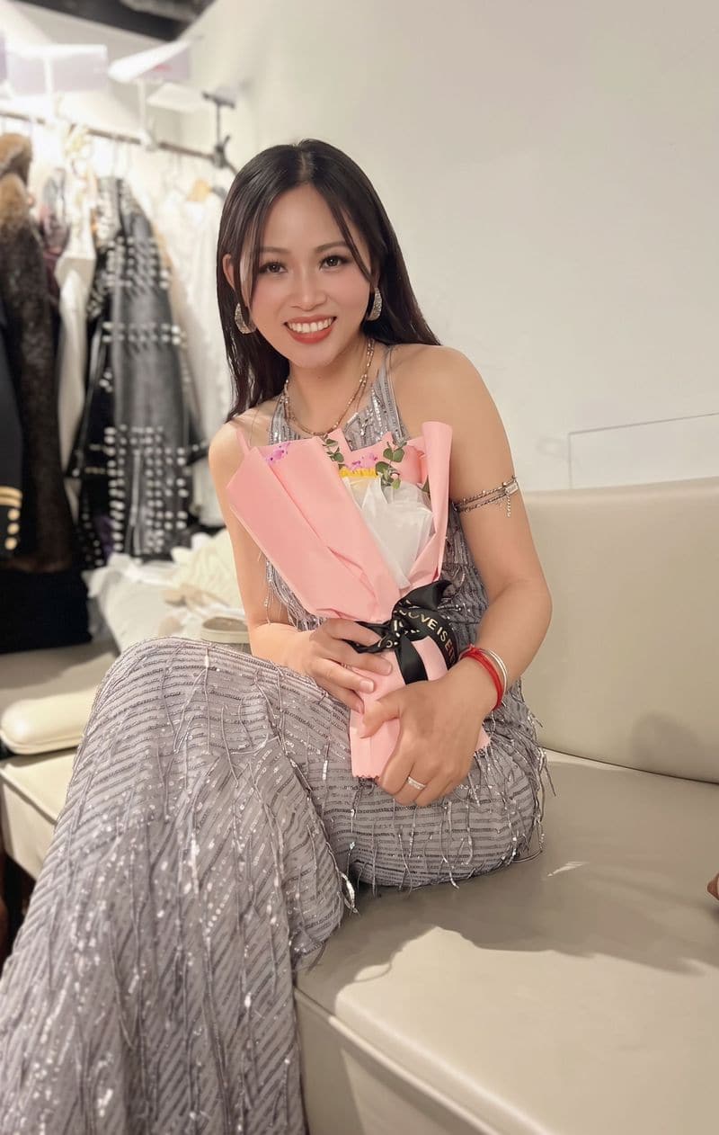 Jaya Lee seated in a dressing room in a silver fringed outfit, holding a pink bouquet