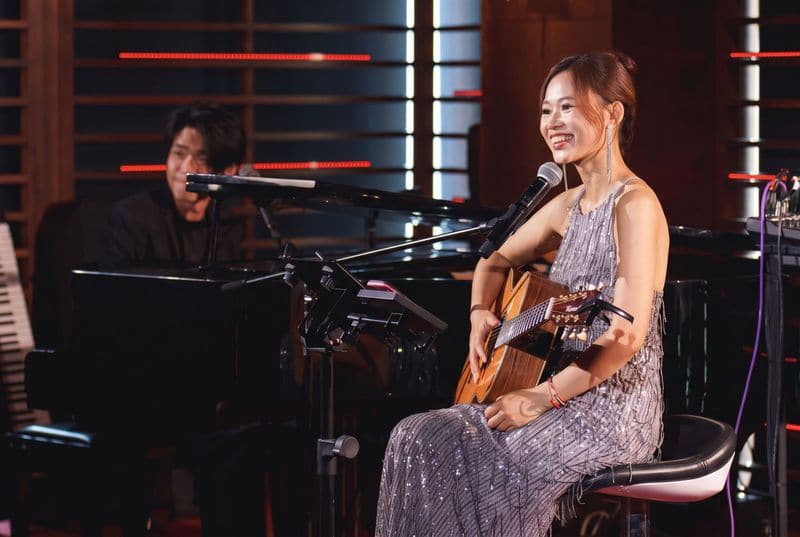Jaya Lee seated with an acoustic guitar, a pianist at a grand piano behind her